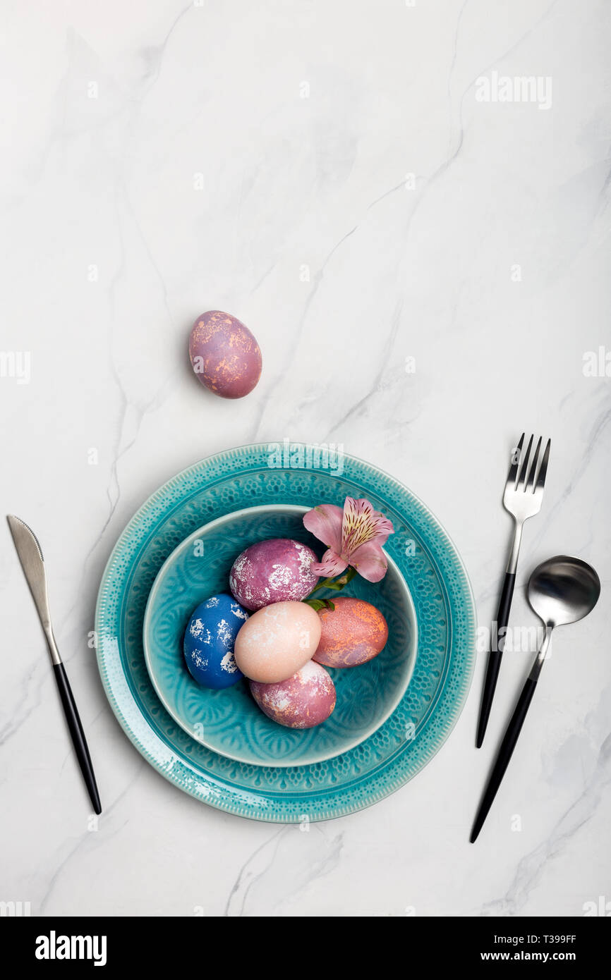 Elegance table setting with colorful Easter eggs in blue plate on white marble background, pink egg near. Concept of Holiday dinner. Flat lay, table t Stock Photo