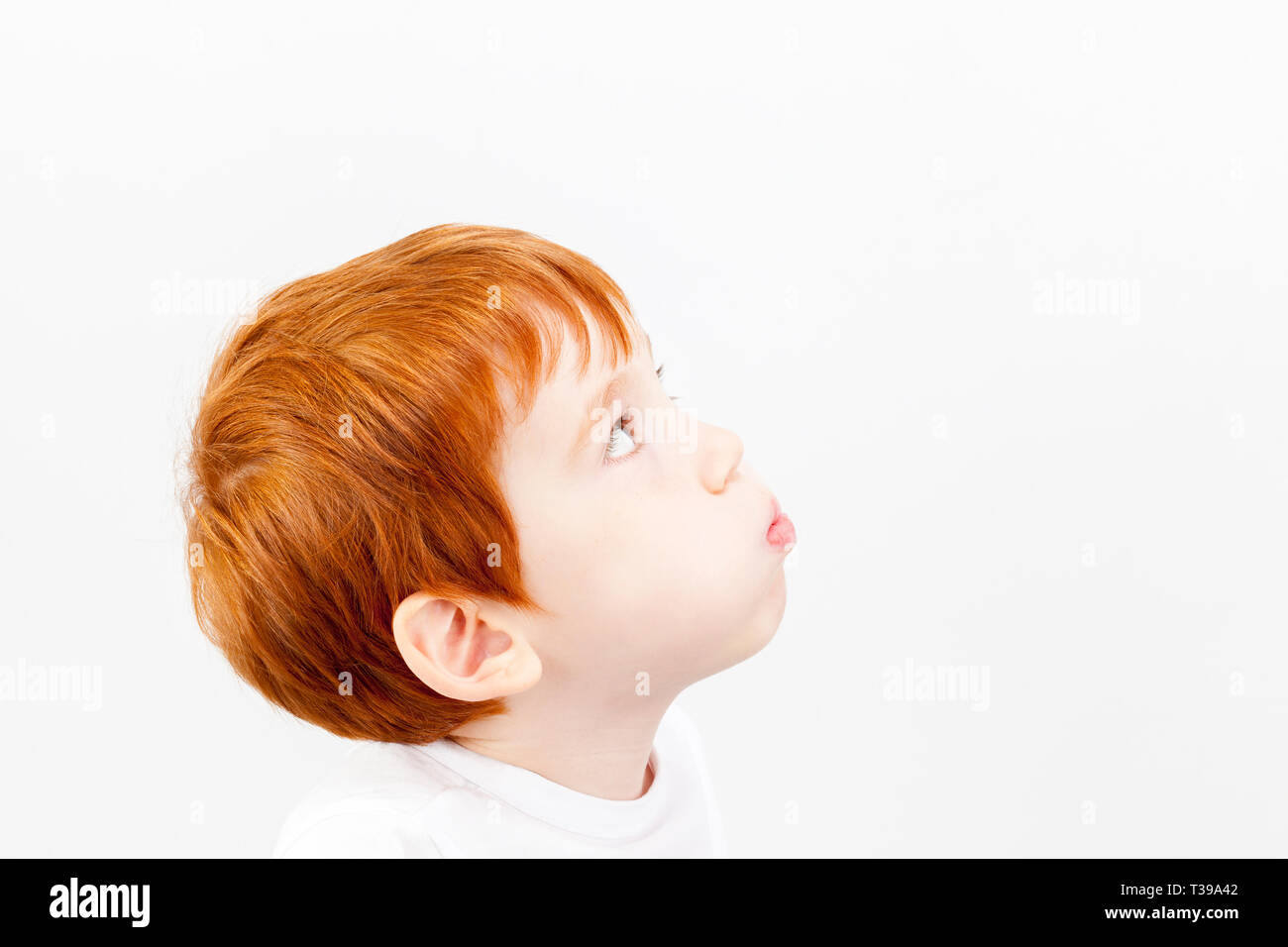 A boy in a white T-shirt with red hair of European appearance looks up, puffed out cheeks Stock Photo