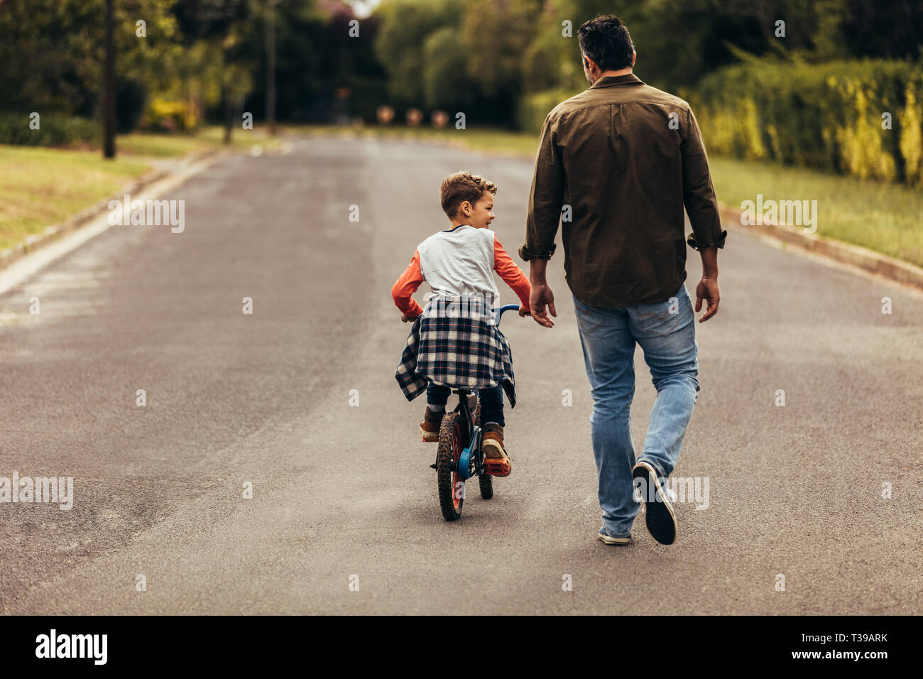kid learning to ride a bicycle on an empty road. Rear view of a boy riding a bicycle while his father walks along with the kid. Stock Photo