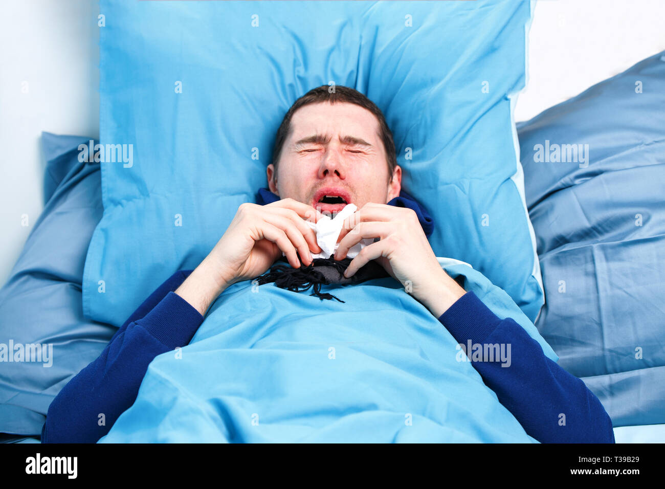 Photo of sneezing sick man in scarf and with handkerchiefs lying in bed. Stock Photo