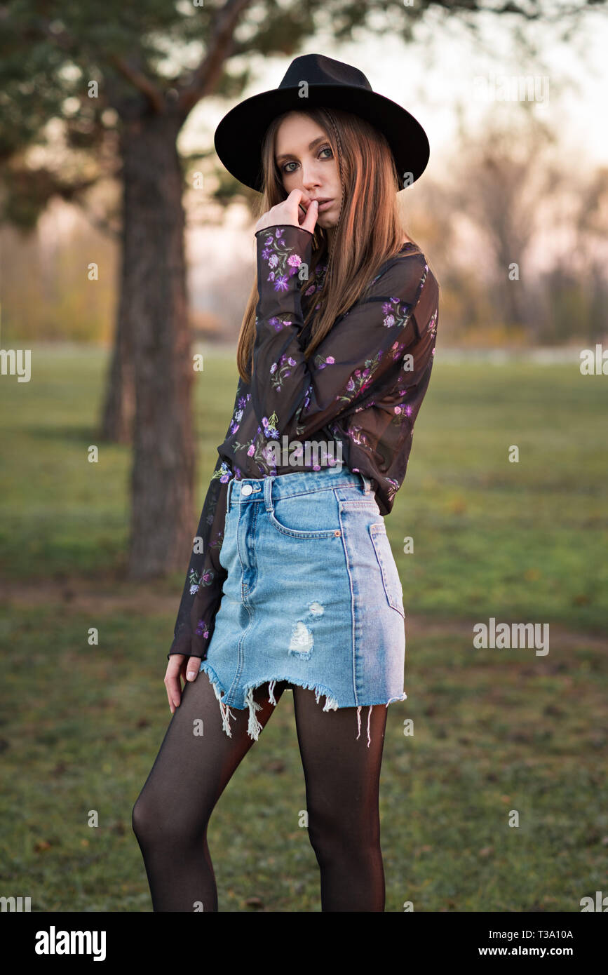 Beautiful brunette in a transparent black blouse, torn denim skirt and hat.  Fashionable woman. Casual wear. Trend clothing Stock Photo - Alamy