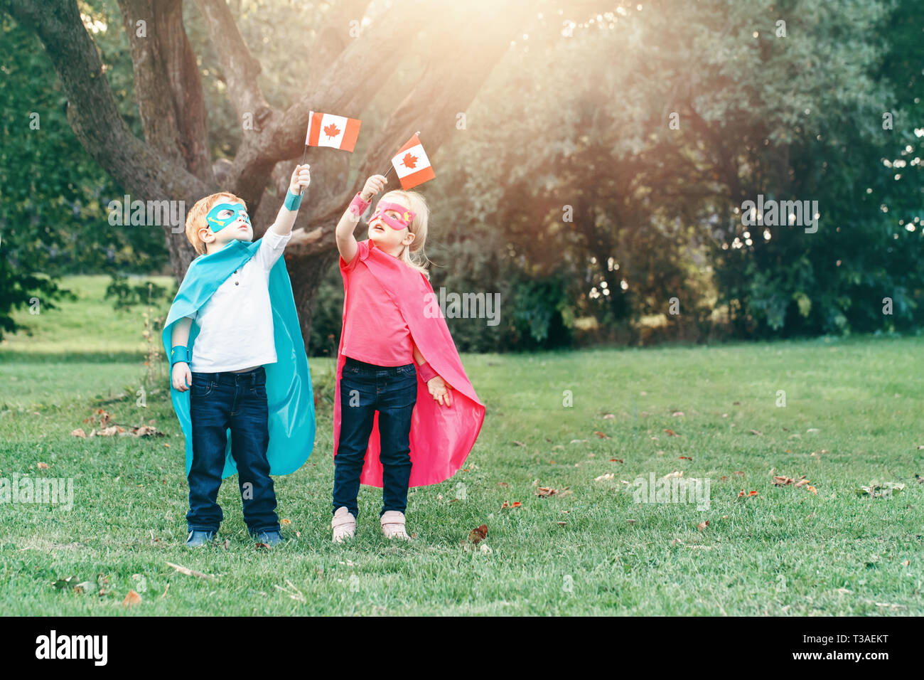 Caucasian children in superhero costumes and masks holding waving Canadian flag. Boys, girl celebrating national holiday Canada day in summer park Stock Photo
