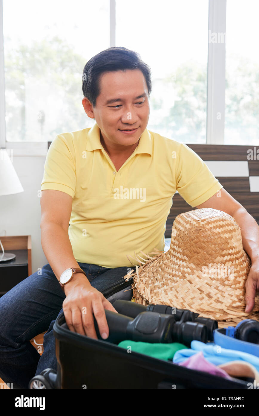 Mature Vietnamese man packing suitcase with hat and binoculars Stock