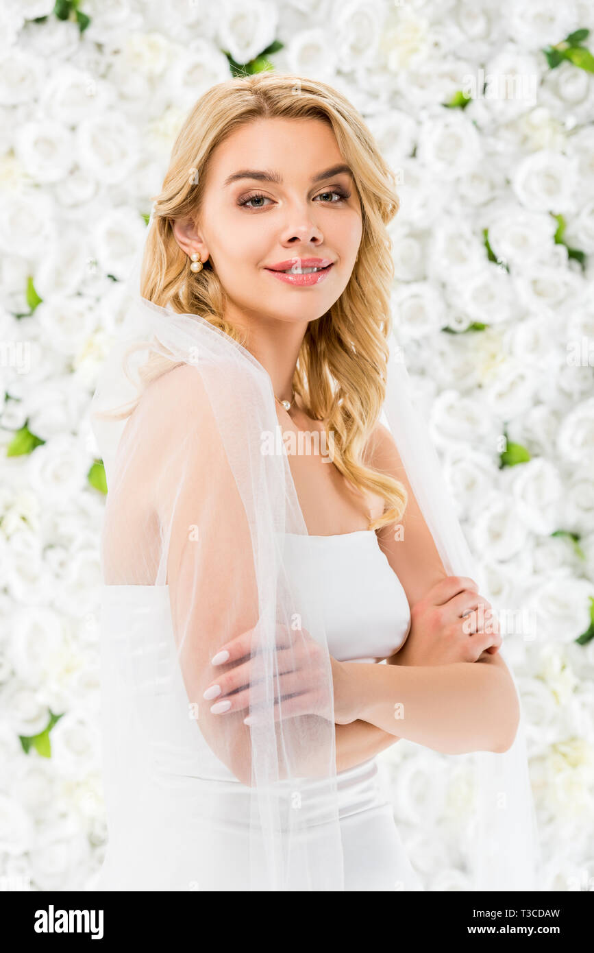 smiling beautiful bride with crossed hands looking at camera on white floral background Stock Photo