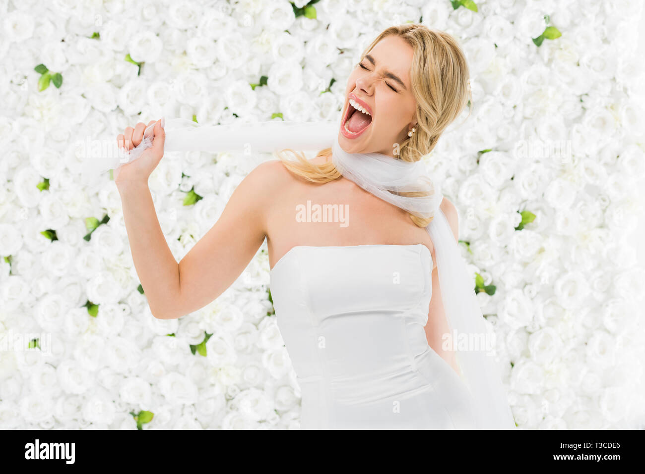 excited young woman winding bridal veil around neck on white floral background Stock Photo