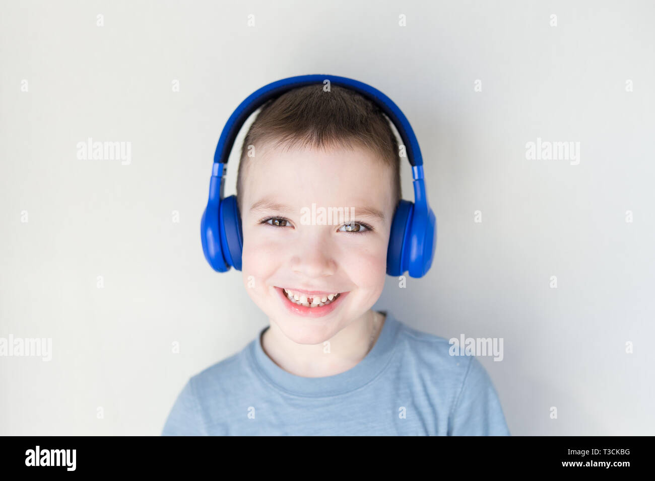 The young boy is smiling and listening to music in blue headphones, standing in front of white wall. Stock Photo