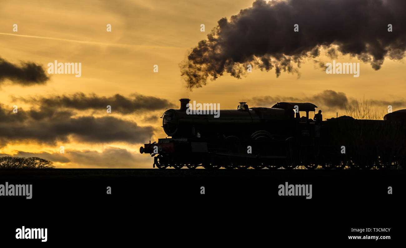 Dramatic, panoramic evening sunset landscape with side view of moving vintage UK steam locomotive in silhouette puffing steam, golden sky background. Stock Photo