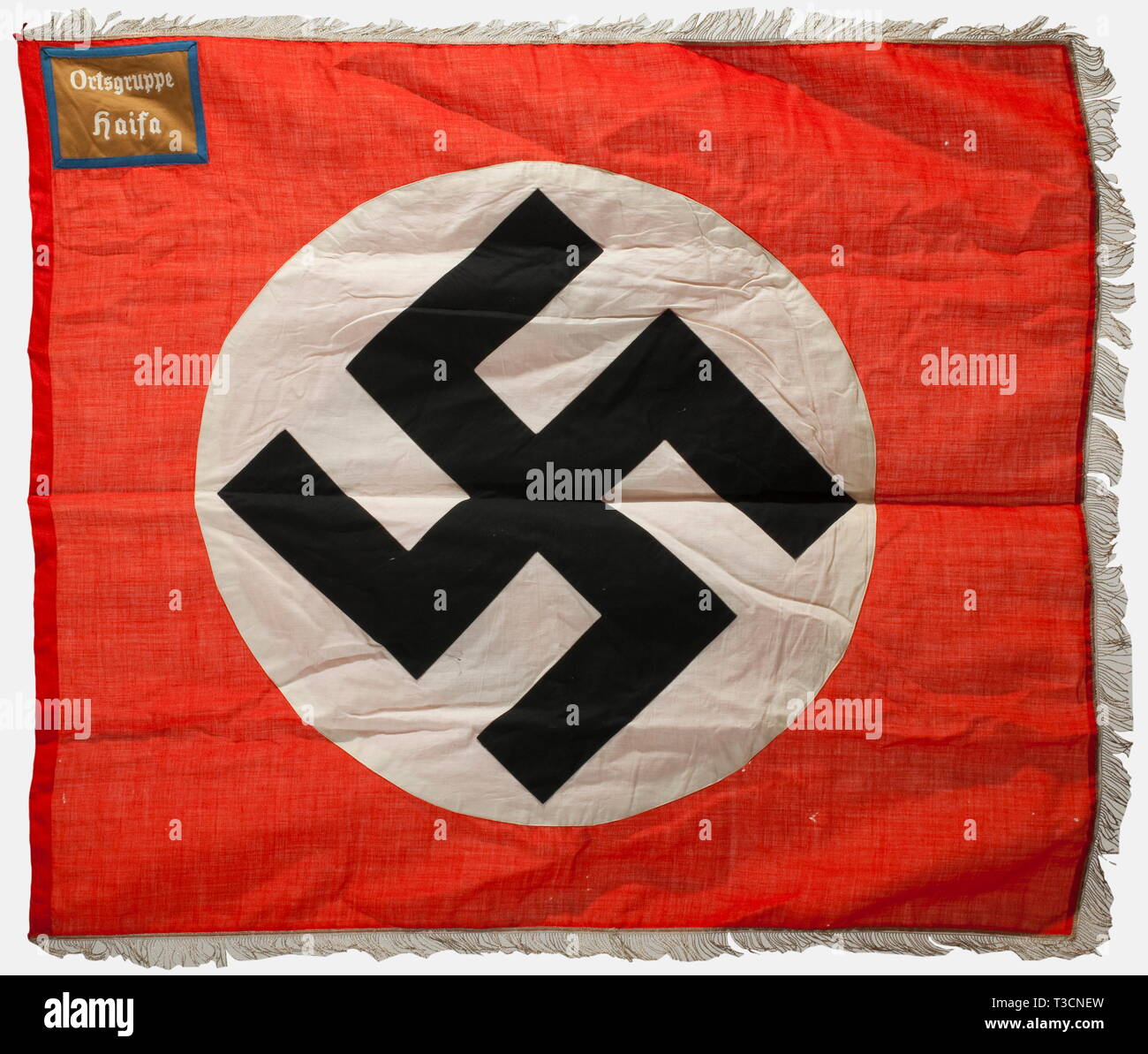 A banner for the Haifa local group, NSDAP Foreign Organization, Palestine National Group Red naval banner cloth with a white disc in the centre on both sides displaying an upright black swastika, a brown panel with blue border trim in the upper hoist corner bearing the white embroidered unit designation, 'Orstgruppe Haifa', silver fringe on three sides, and six nickel-plated flagpole rings, the top one missing. Small holes, generally in very good condition. Dimensions 125 x 135 cm. The NSDAP Haifa local group was part of the Palestine National Group of the NSDAP, whose memb, Editorial-Use-Only Stock Photo