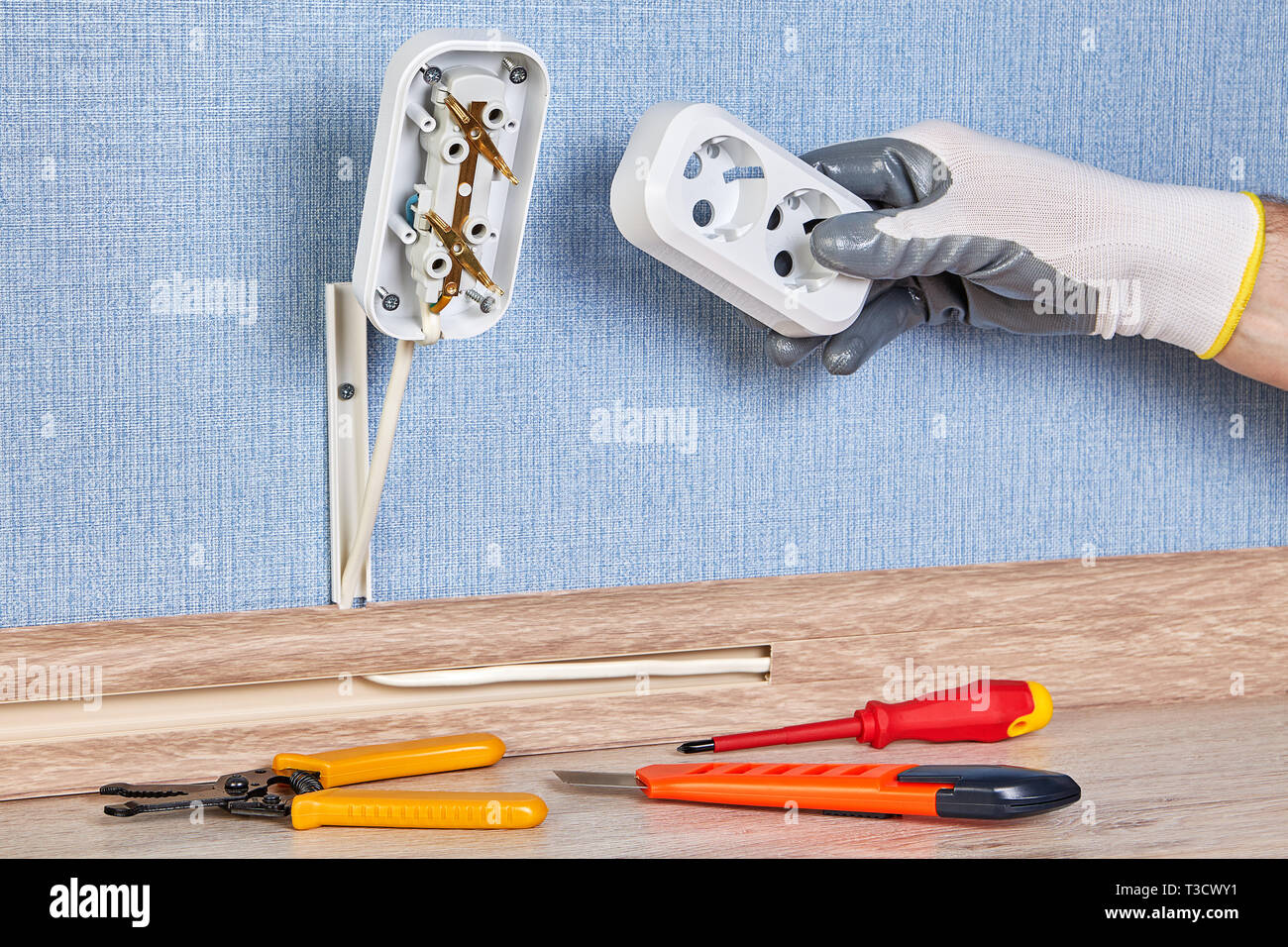 Worker in protective gloves is installing wall plug socket, there are hand tools on the floor. Stock Photo