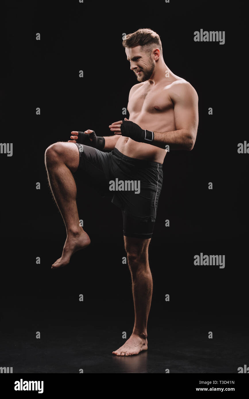 muscular barefoot mma fighter in bandages doing kick with knee on black