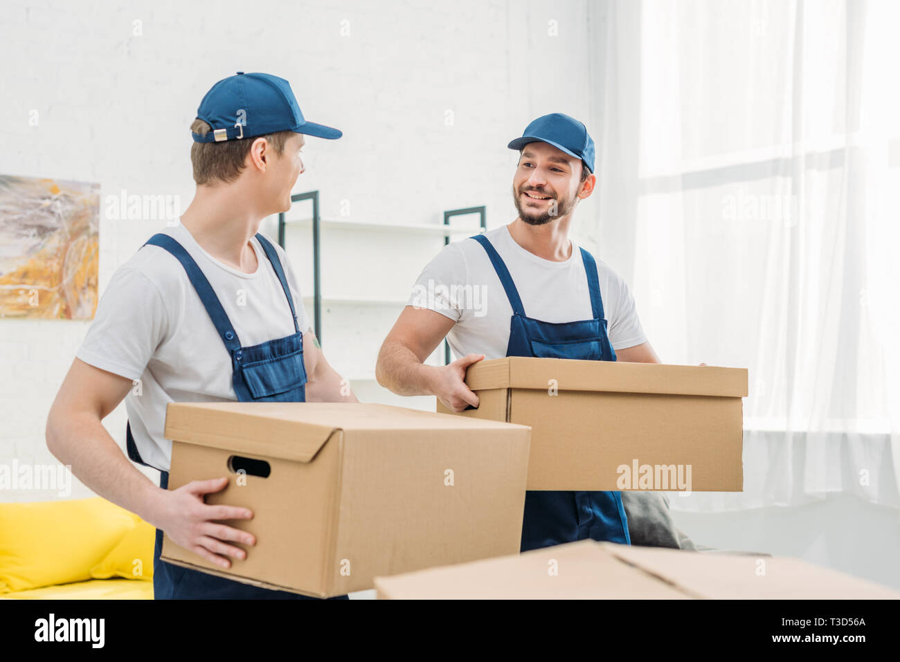two smiling movers looking at each other while transporting cardboard boxes in apartment Stock Photo