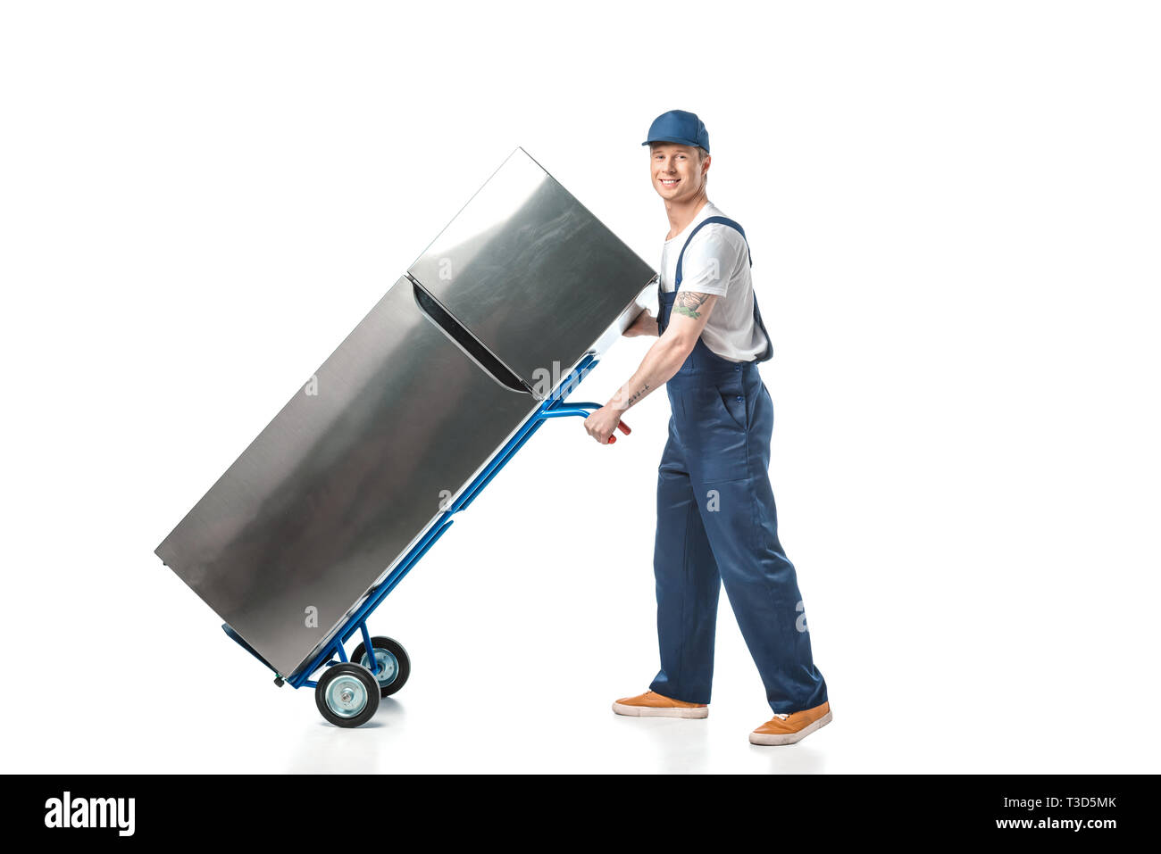 handsome mover in uniform looking at camera and transporting refrigerator on hand truck isolated on white Stock Photo