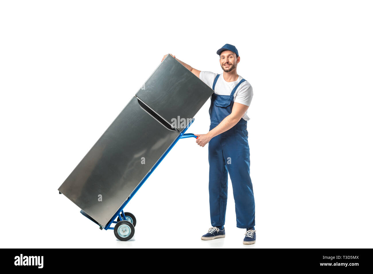 handsome mover in uniform looking at camera and transporting refrigerator on hand truck isolated on white Stock Photo