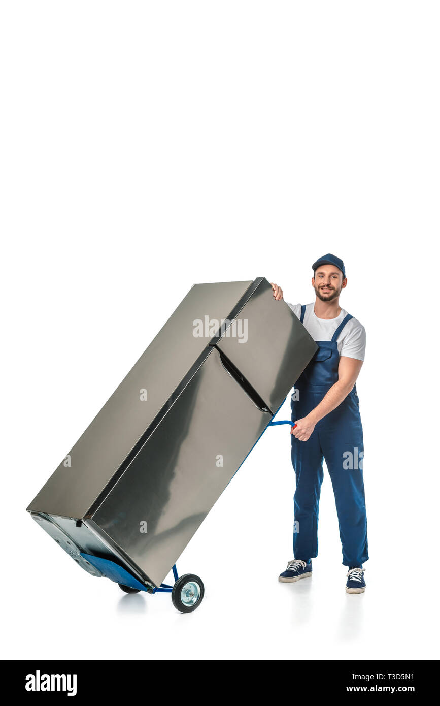handsome mover in unifrom looking at camera and transporting refrigerator on hand truck isolated on white with copy space Stock Photo