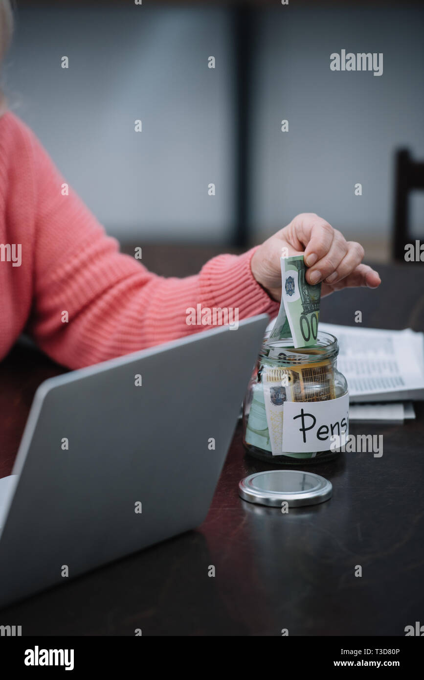 cropped view of senior woman sitting at table with laptop while putting money in glass jar with 'pension' word Stock Photo