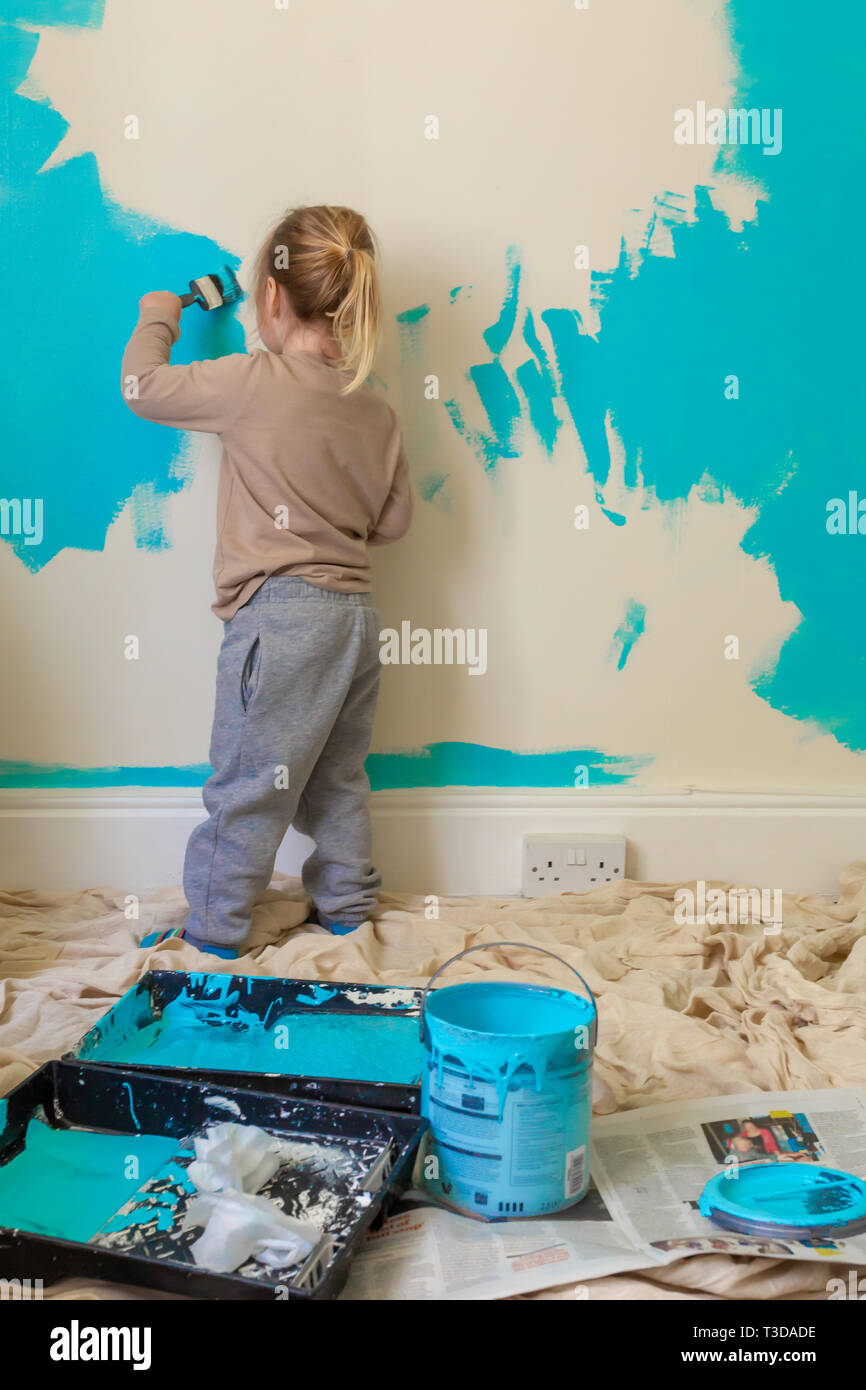 Candid colour photograph of young child painting white wall blue with back to camera. Stock Photo