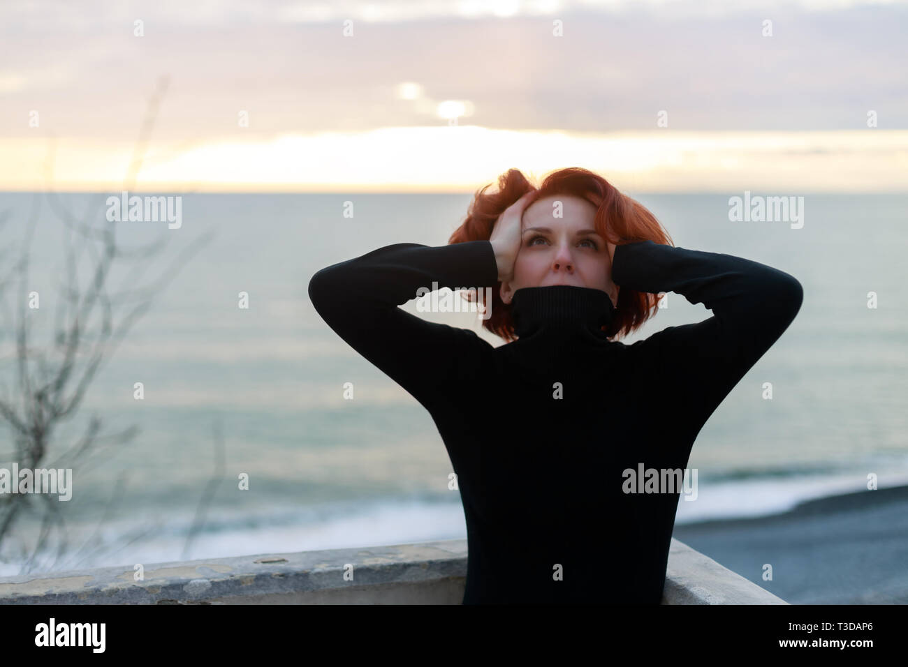 Young woman, in despair, put her hands on her head, directing her gaze towards God. Portrait of a woman with red hair against the background of the se Stock Photo