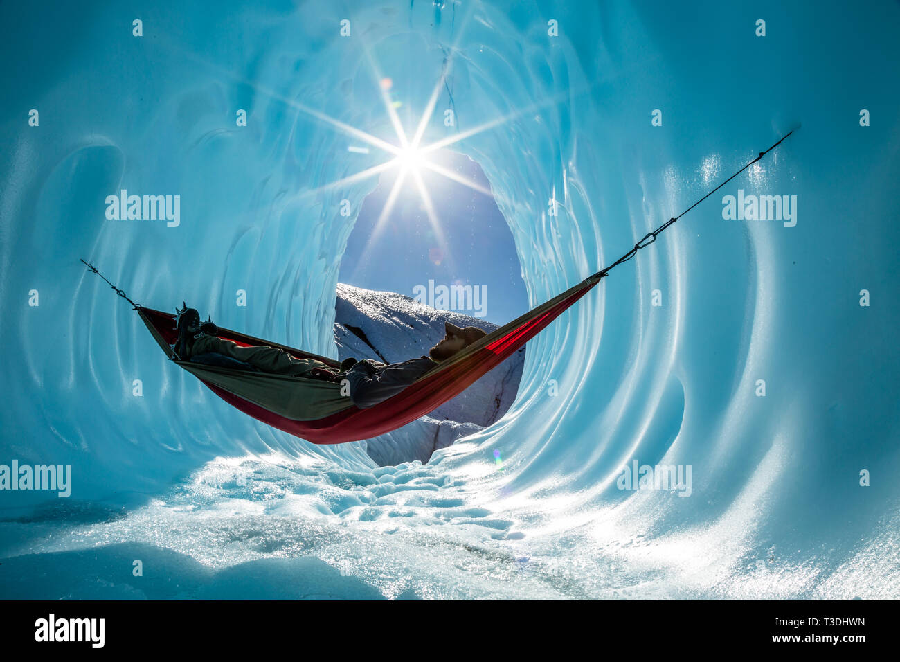 In the wilderness of Alaska, an ice climber has set up a hammock to rest inside of an ice cave on the Matanuska Glacier. The sun peeks in from above Stock Photo