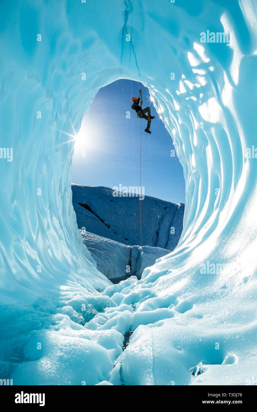 Hanging from a rope fixed above the entrance of a large blue ice cave, an ice climber ascends out of the glacial cavern. The scene is from the Matanus Stock Photo