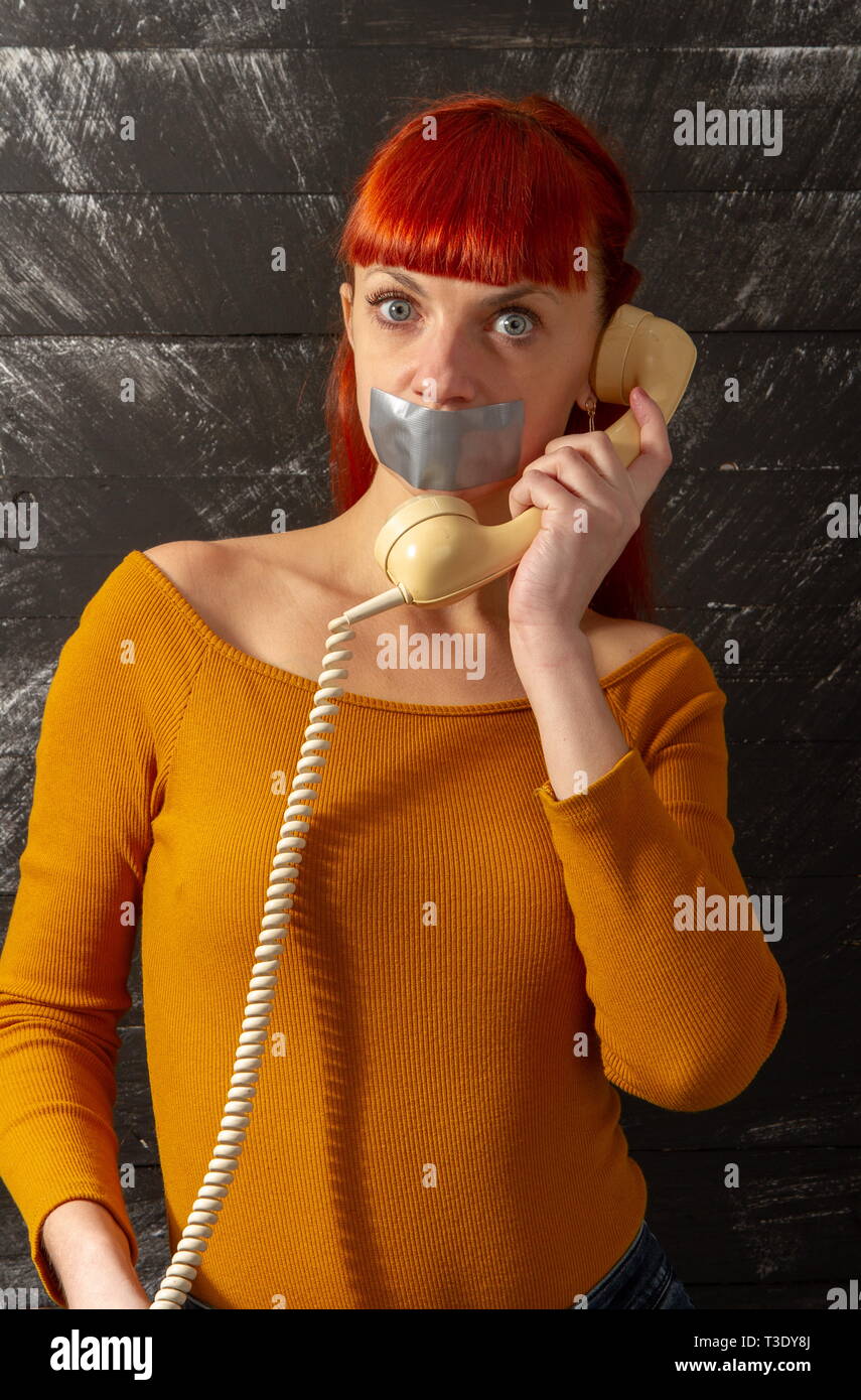 Conceptual picture. Young red-haired girl tries to talk on the phone with sealed with adhesive tape with her lips so that she keeps silence Stock Photo