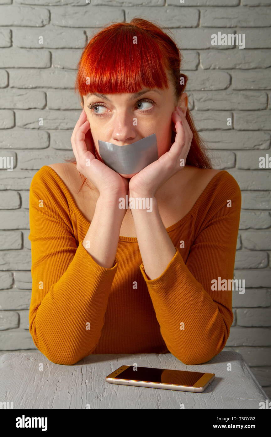 Conceptual picture. Young red-haired girl sitting near a smartphone with adhesive tape lips to keep silence Stock Photo