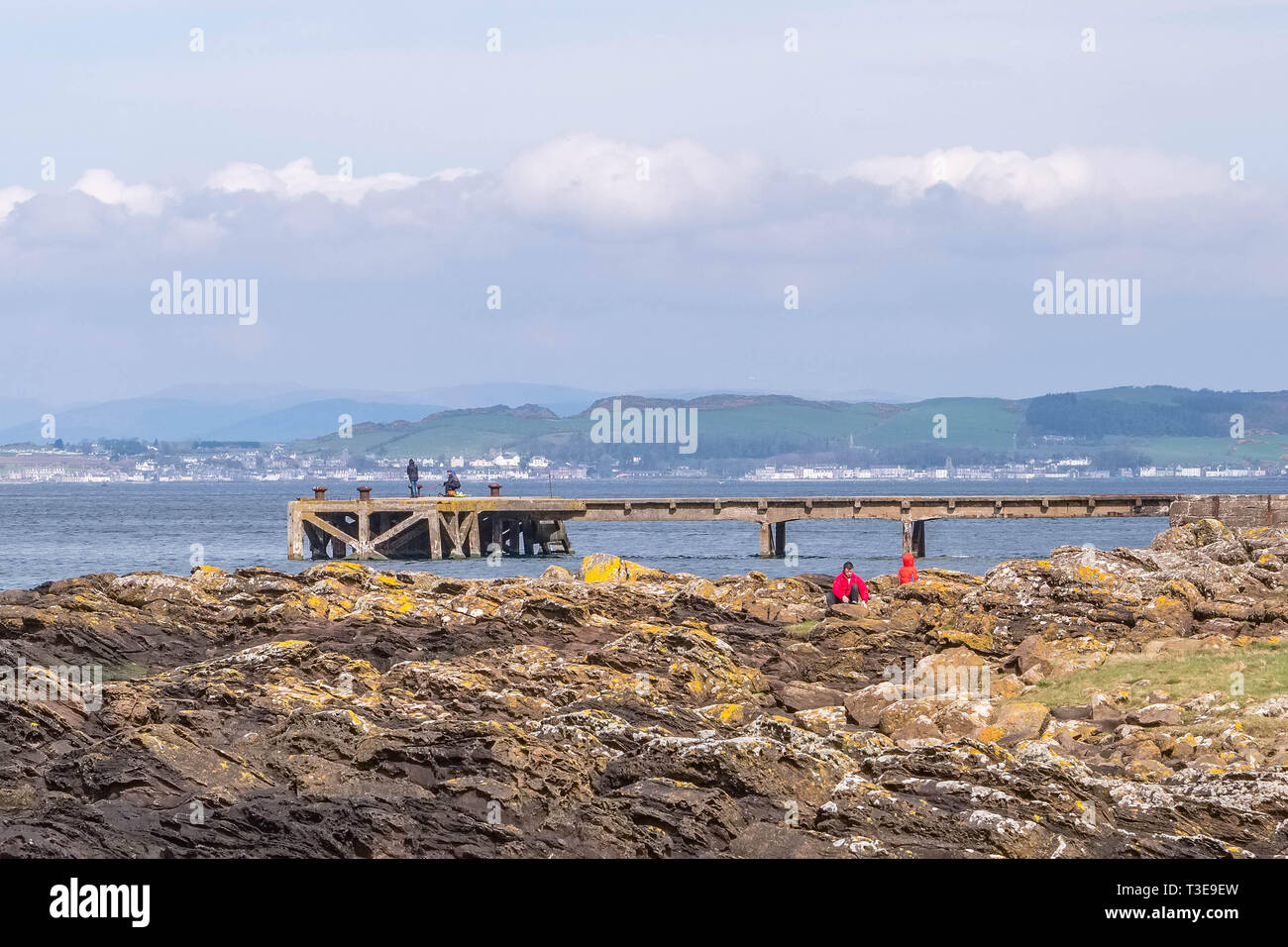 Seamill, Scotland, UK - April 05, 2019: Some people down amongst the jagged lichen coverted rocks and fishermen on the old Seamill jetty. Stock Photo