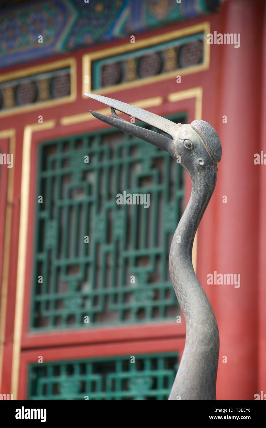 Silver coloured Chinese sculpture of a stork with bendy neck, partially open beak and a hat. Green red and gold oriental building in background. Stock Photo