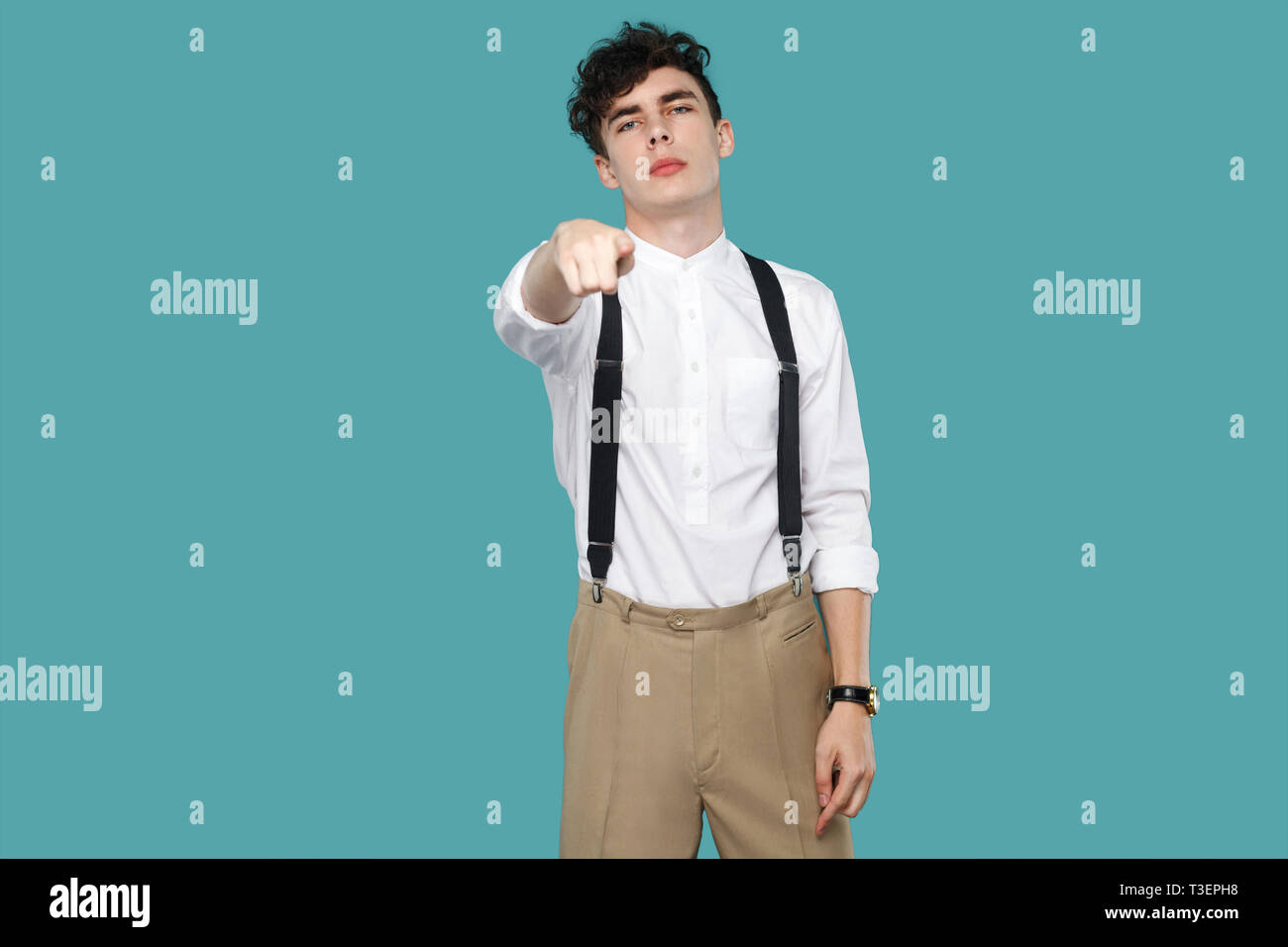 Serious man pointing and looking at camera. Portrait of handsome hipster curly young businessman in classic casual white shirt and suspender standing. Stock Photo