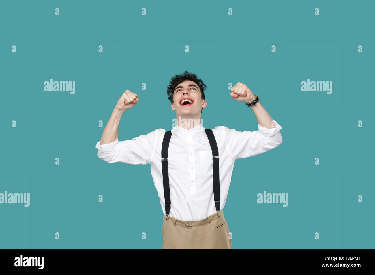 Happy excited man celebrating his victory. Portrait of handsome hipster curly young businessman in classic casual white shirt and suspender standing.  Stock Photo