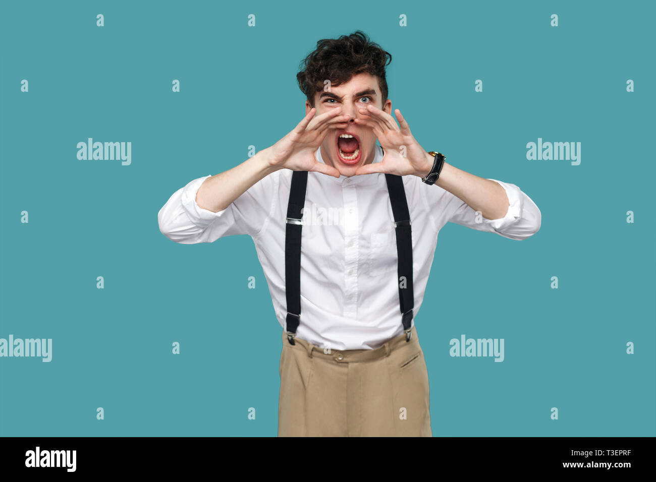 Angry man screaming and looking at camera. Portrait of handsome hipster curly young businessman in classic casual white shirt and suspender standing.  Stock Photo