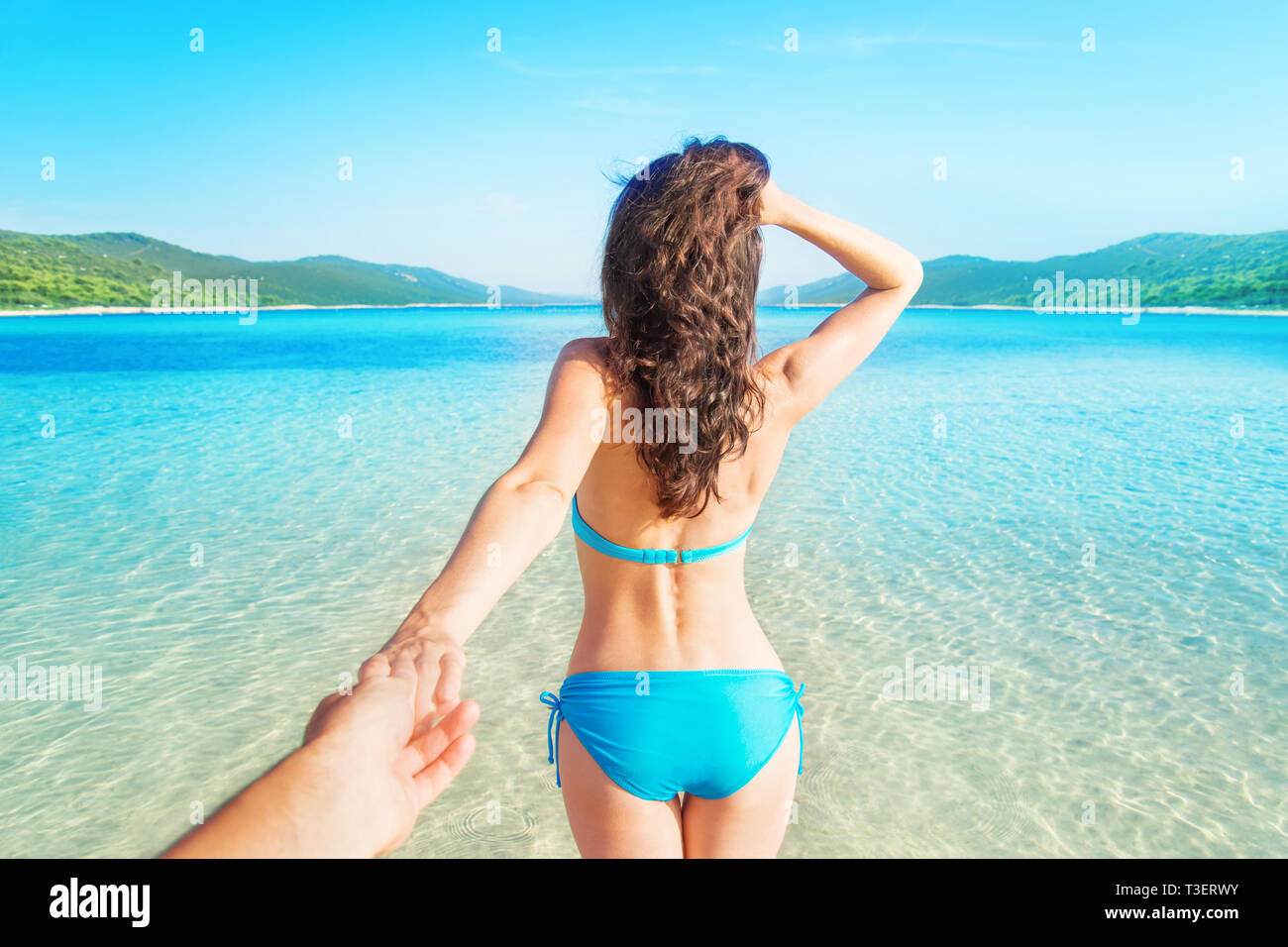 young girl wearing blue swimsuit with her back to camera leading to sea with her hand on empty beautiful beach with clear water Stock Photo