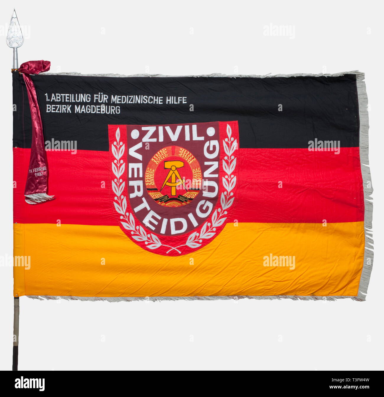 A banner for the GDR civil defense, 1st Medical Assistance Detachment, Magdeburg District Banner cloth of a black/red/gold mixed weave. Dimensions 183 x 117 cm. It displays the machine-embroidered civil defense emblem in the centre and the unit identification at the top hoist. Silver grey fringe. On a two piece flagstaff with an aluminium finial and a wine red artificial silk flag ribbon inscribed '11 February 1987'. It comes with the protective sleeve for the banner. historic, historical, 1980s, 20th century, GDR, East Germany, Eastern Germany, , Additional-Rights-Clearance-Info-Not-Available Stock Photo