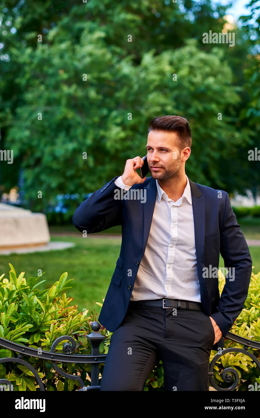 A handsome young businessman leaning against the fence in a park while talking on a phone Stock Photo