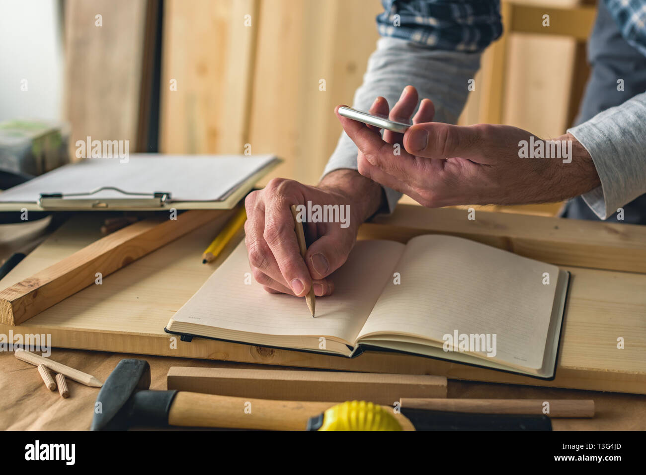 Carpenter is writing project notes while using smartphone in small business woodwork workshop Stock Photo