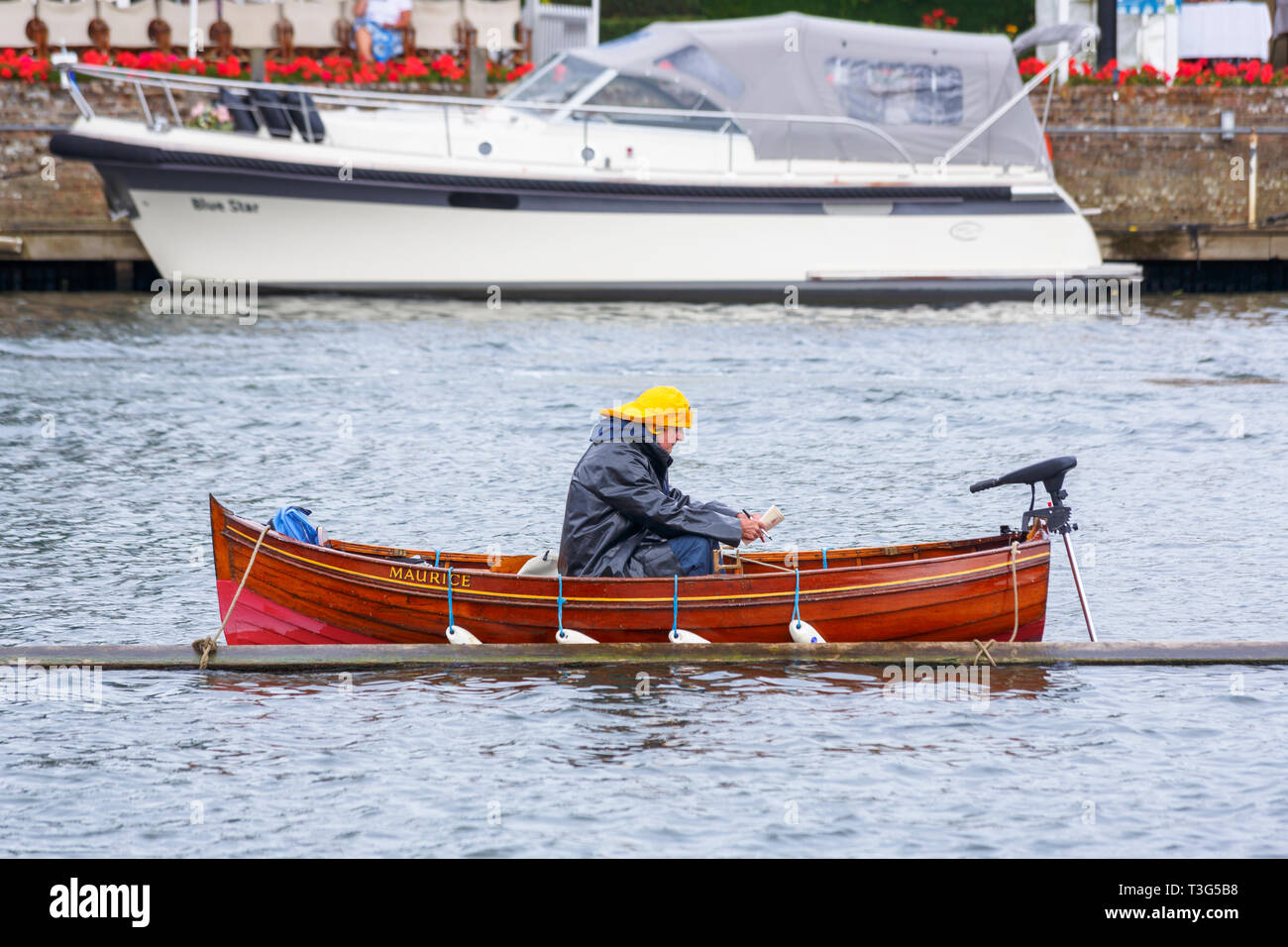 A steward wearing a raincoat and yellow sou'wester hat sits in a wooden rowing boat moored in the River Thames on a wet day at Henley Royal Regatta Stock Photo
