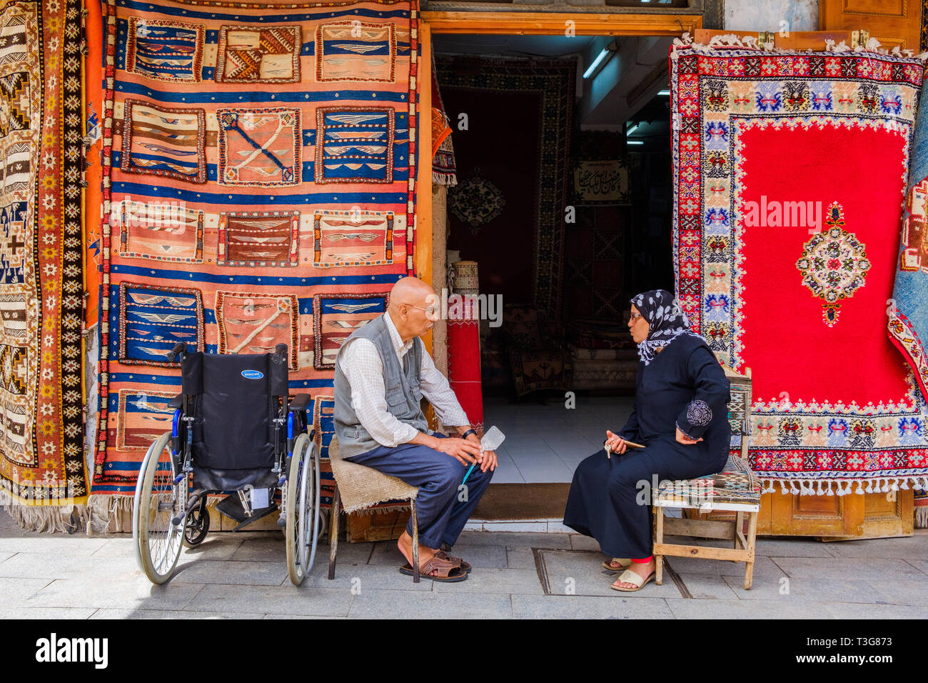 Morocco, Rabat. Lanes and shops in the medina. Man and woman sitting on chairs, talking in front of the shop of a carpet seller, in the street “rue de Stock Photo
