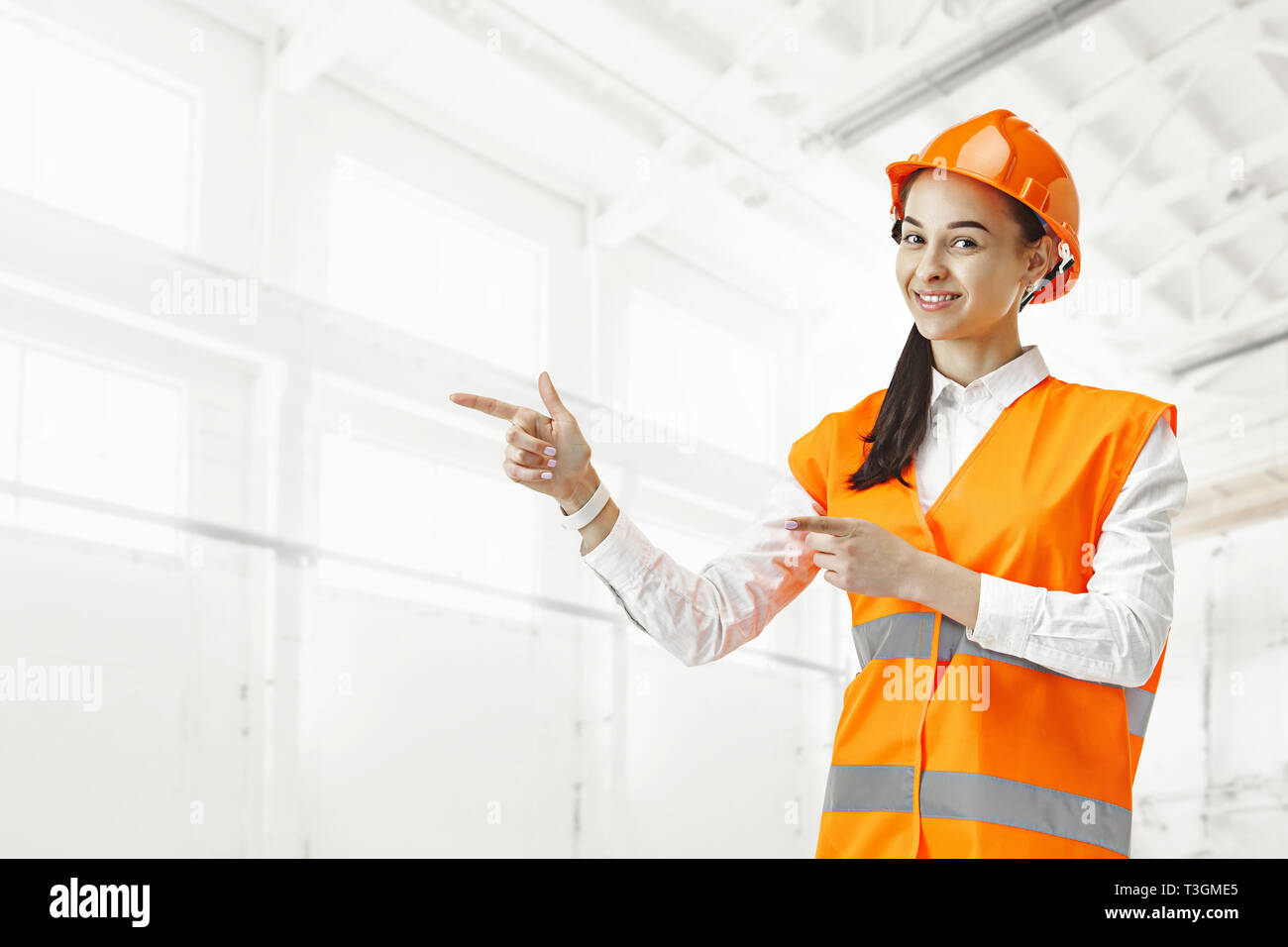 Destroying gender stereotypes. Female builder in male profession. Woman standing against industrial background and pointing to left. Safety specialist, engineer, occupation, businesswoman, job concept. Stock Photo