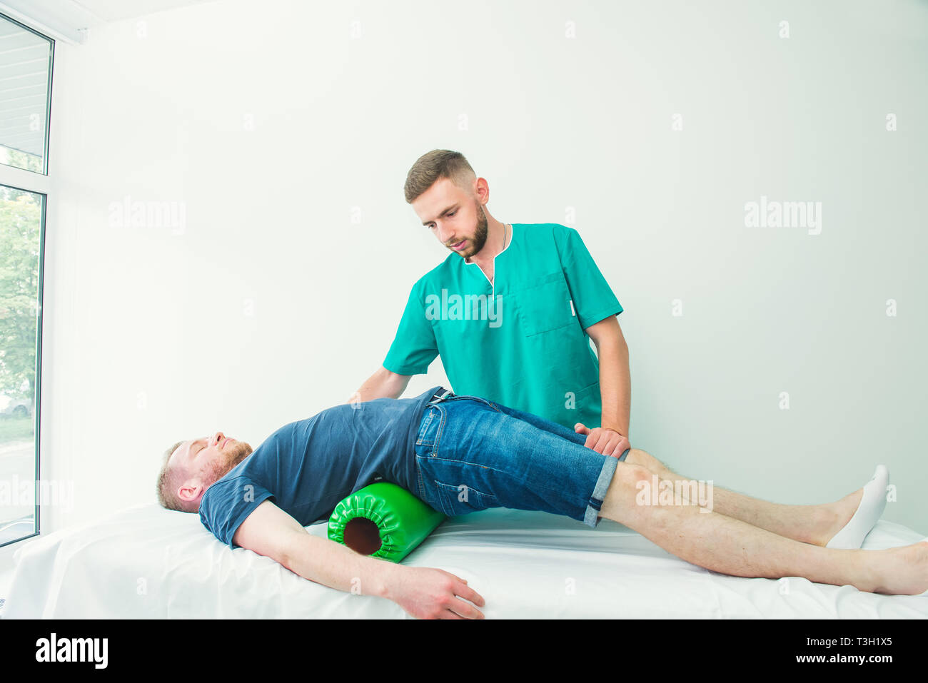 Man at the physiotherapy doing physical exercises with his therapist, they using a massage roll. A chiropractor treats patient's loins spine in medica Stock Photo