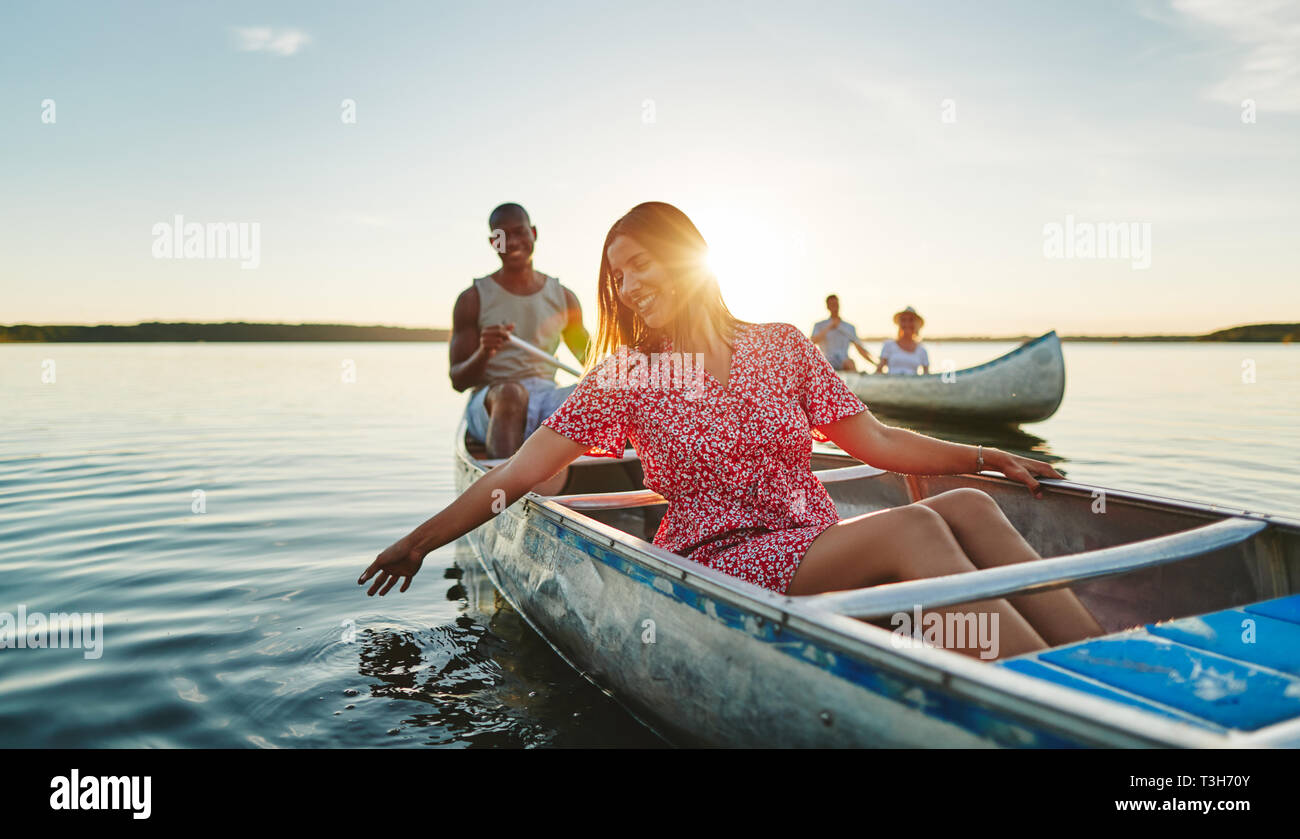 Smiling young woman dipping her hand in lake water while paddling a canoe with her boyfriend and friends on a late summer afternoon Stock Photo