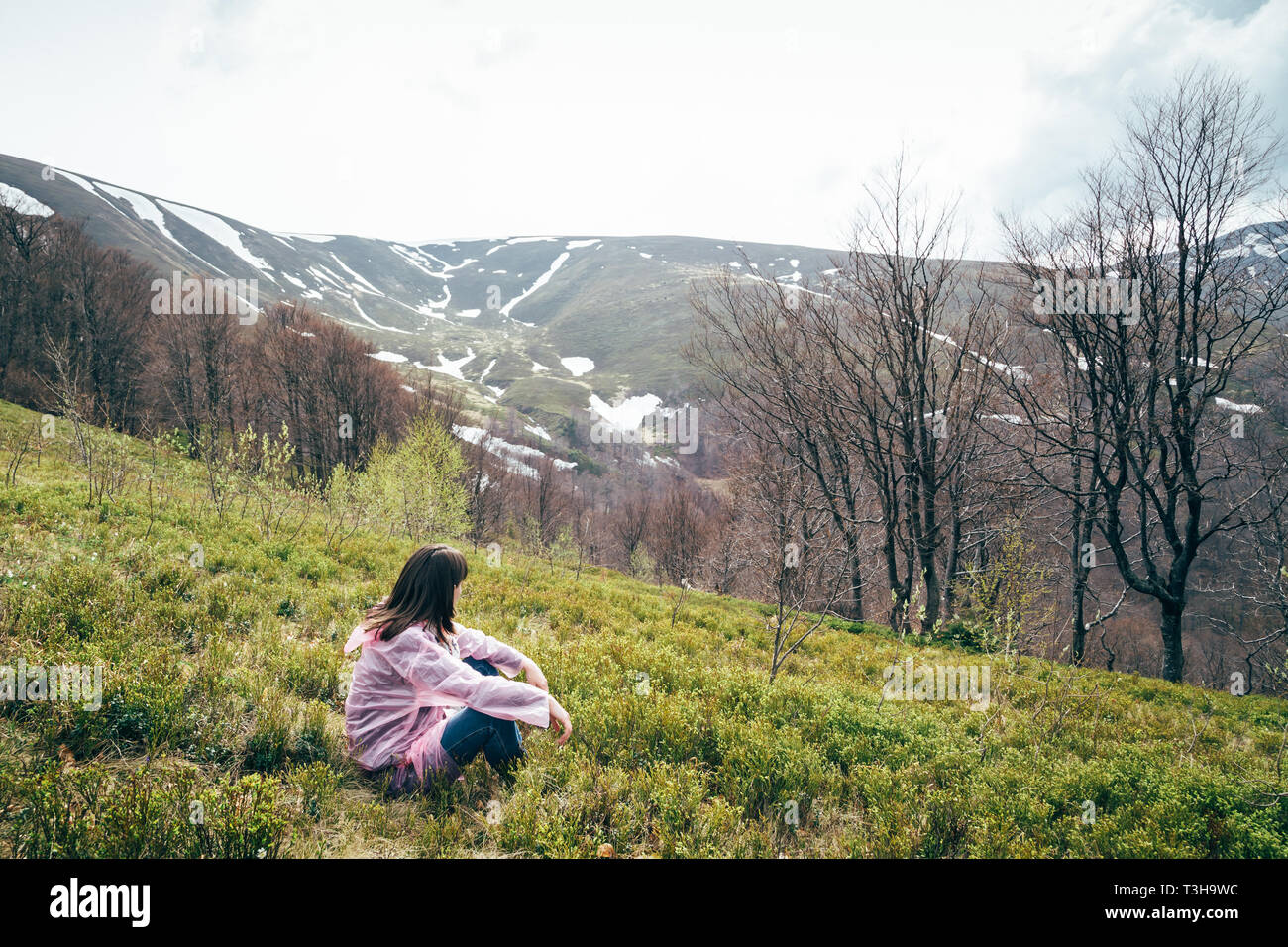 Tourist girl in pink rain jacket sitting on the grass looking at mountains surrounded by forest, enjoying silence and harmony of nature Stock Photo