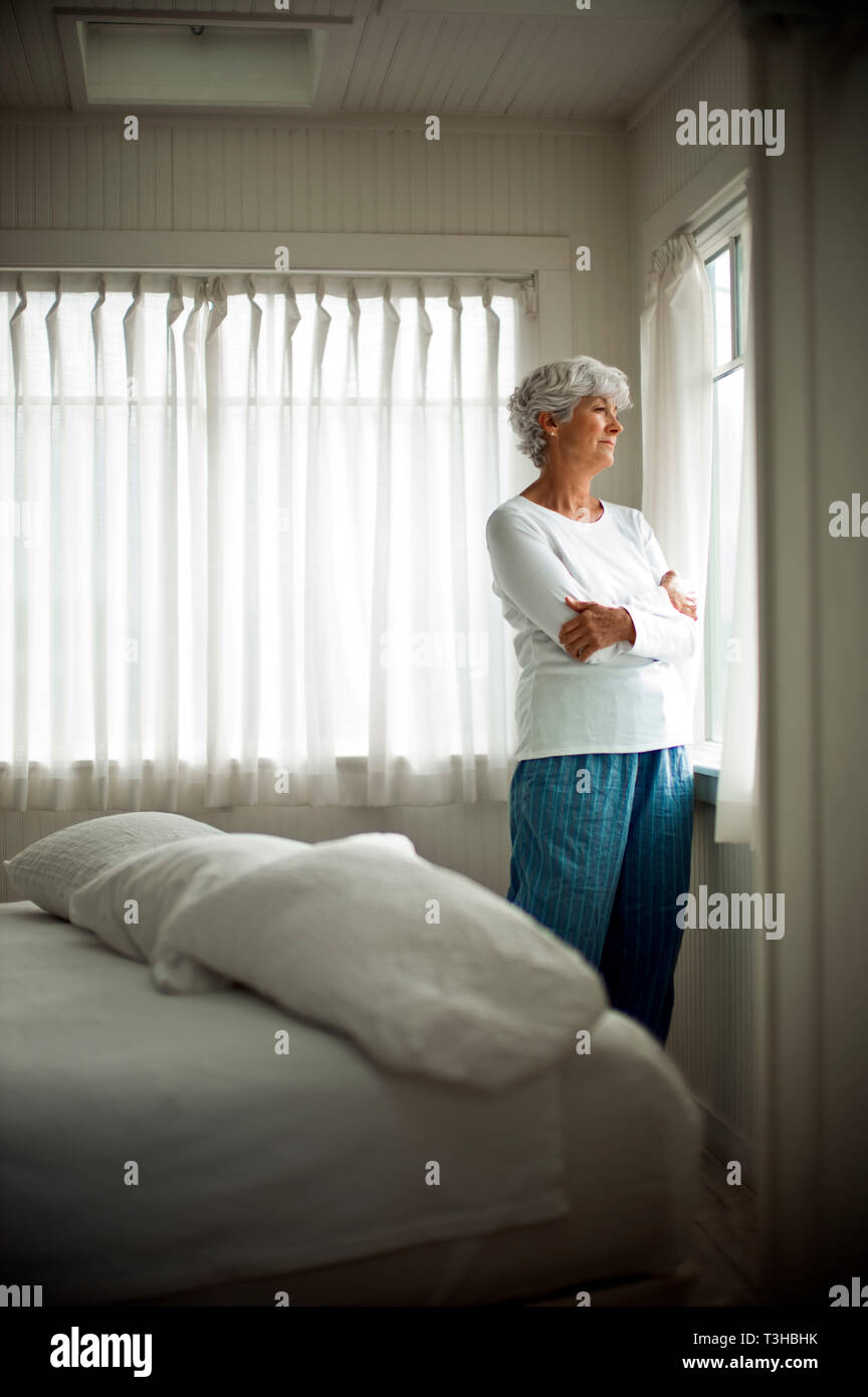 Unhappy mature woman in her pajamas stands with her arms crossed as she looks out of a bedroom window. Stock Photo