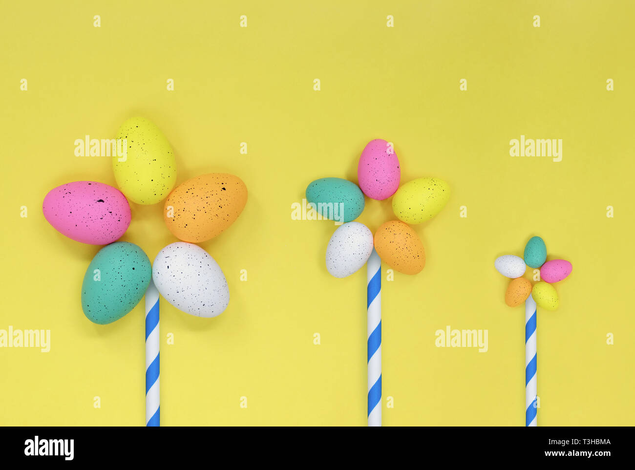 Three flowers made from colourful easter eggs. Flat lay against a yellow background, with space for copy. Stock Photo