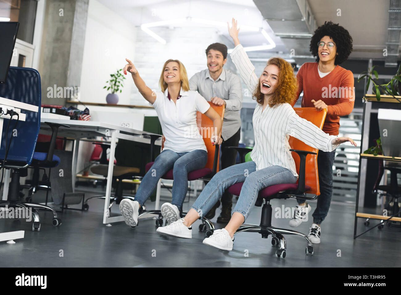 Cheerful millennials riding on office furniture, having fun at work Stock Photo