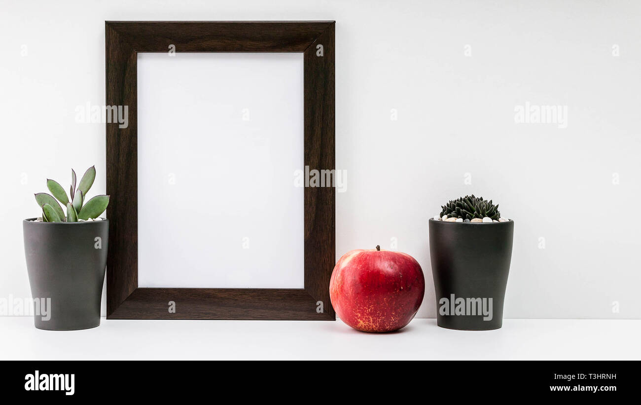 Empty dark photo frame, two succulents in dark pots and a red juicy apple on a white background. Scandinavian style in the interior. Stock Photo