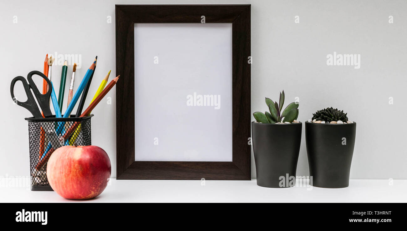 Empty dark photo frame, succulents in dark pots, colored pencils in a stand and a red apple on a white background. MockUp. Scandinavian style in the i Stock Photo