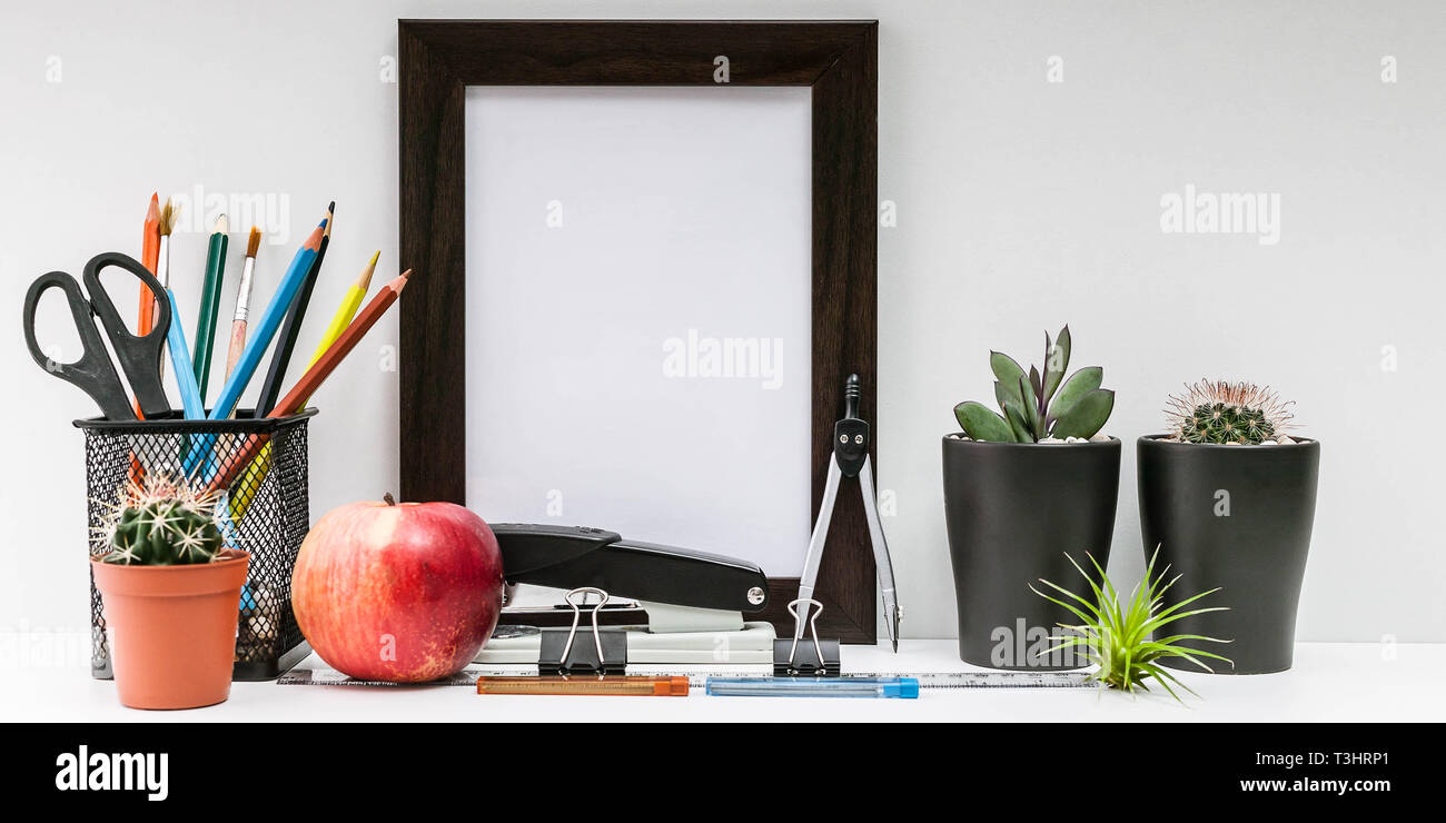 Empty dark photo frame, succulents in dark pots, colored pencils, office supplies, red apple on a white background. Desktop MockUp. Scandinavian style Stock Photo