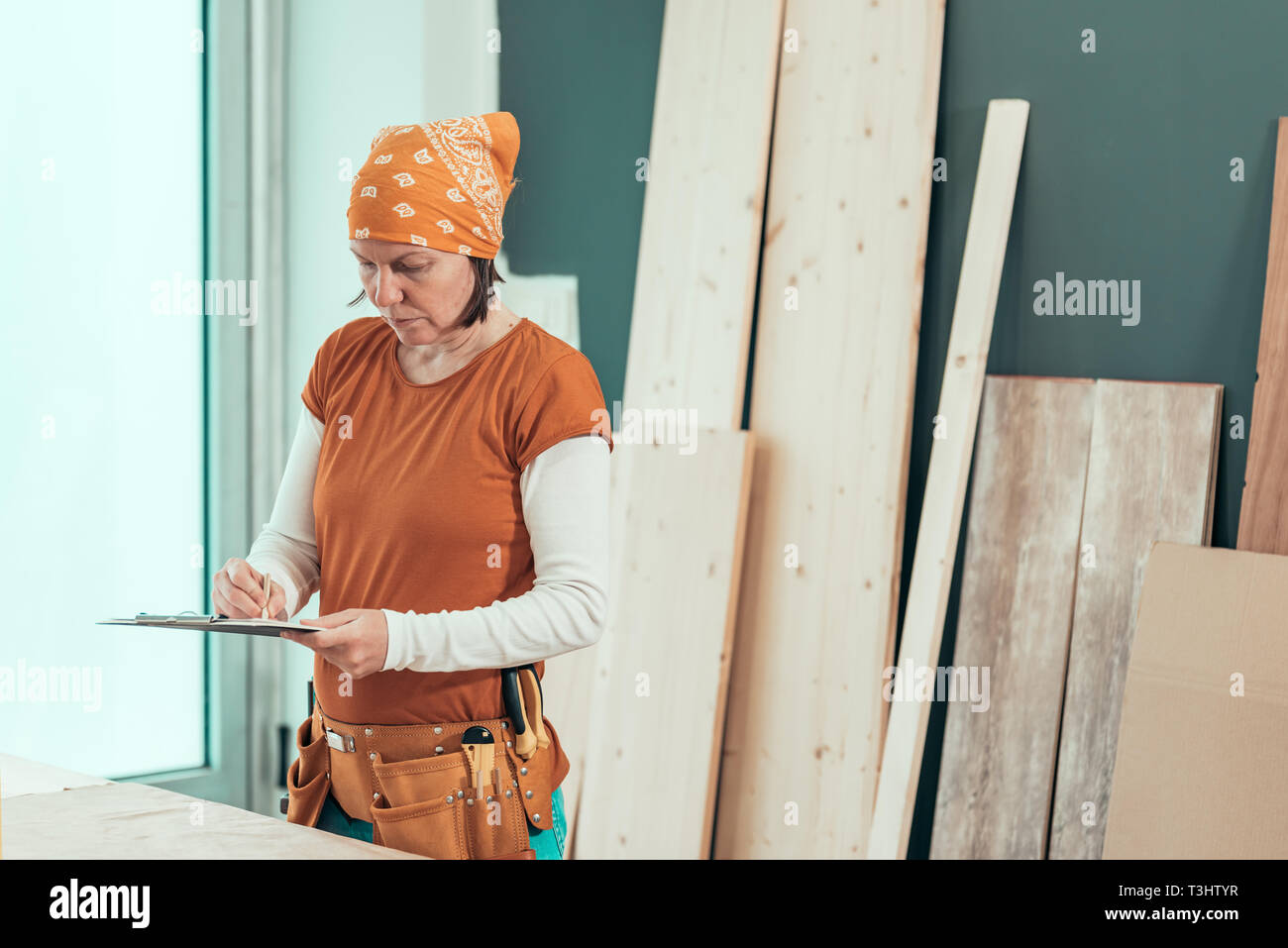 Female carpenter wearing bandanna is writing project notes on clipboard note paper in her small business woodwork workshop Stock Photo