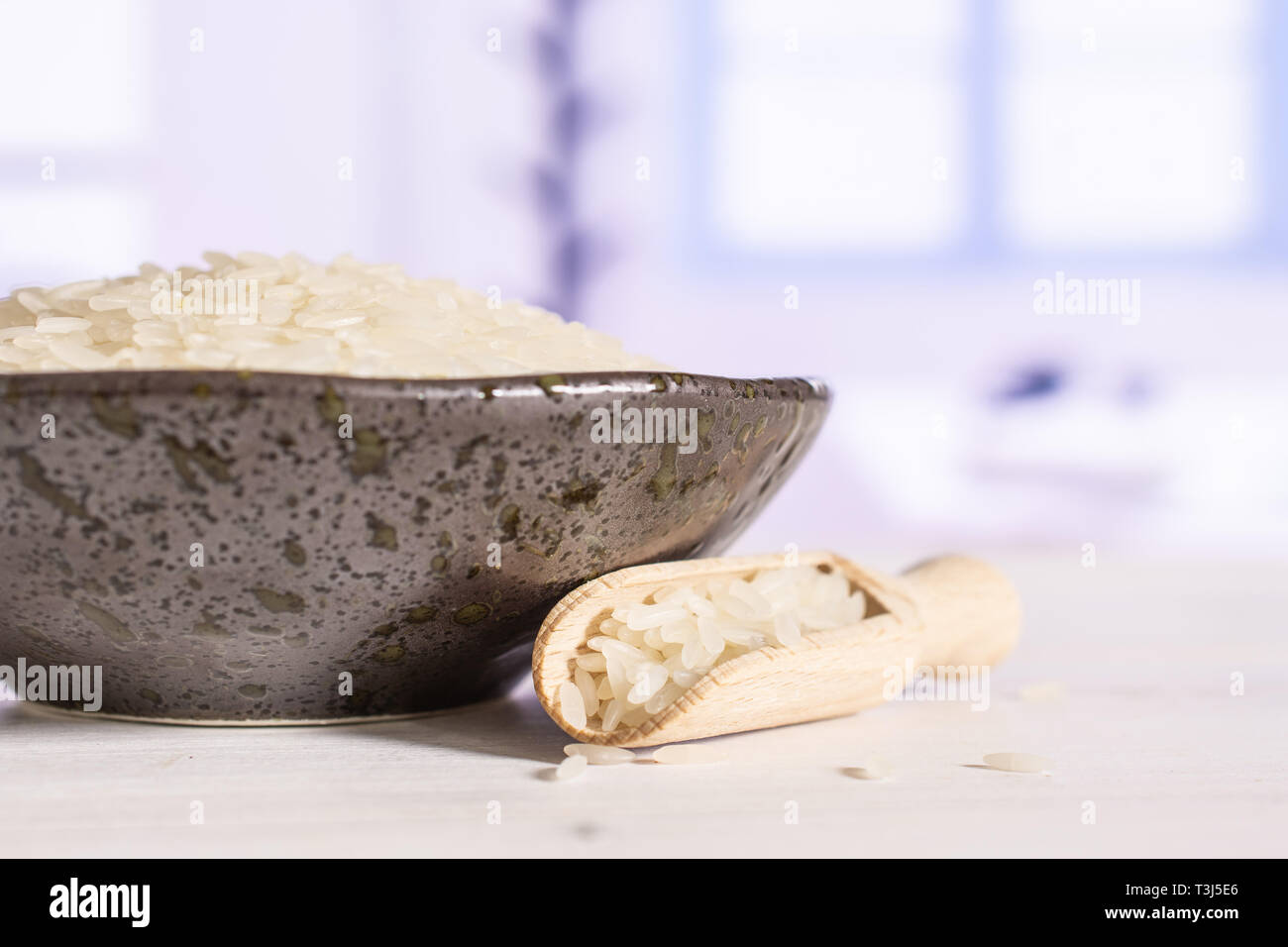 Lot of whole white jasmine rice grains on grey ceramic plate with wooden scoop with blue window in a white kitchen Stock Photo