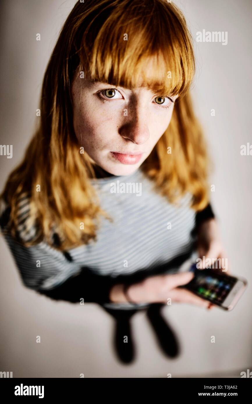 Girl, teenager, red-haired, with smartphone in hand, looks skeptical in camera, studio shot, Germany Stock Photo