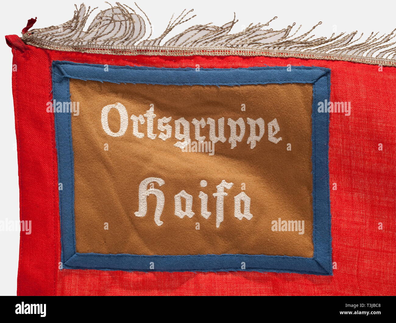 A banner for the Haifa local group, NSDAP Foreign Organization, Palestine National Group Red naval banner cloth with a white disc in the centre on both sides displaying an upright black swastika, a brown panel with blue border trim in the upper hoist corner bearing the white embroidered unit designation, 'Orstgruppe Haifa', silver fringe on three sides, and six nickel-plated flagpole rings, the top one missing. Small holes, generally in very good condition. Dimensions 125 x 135 cm. The NSDAP Haifa local group was part of the Palestine National Group of the NSDAP, whose memb, Editorial-Use-Only Stock Photo